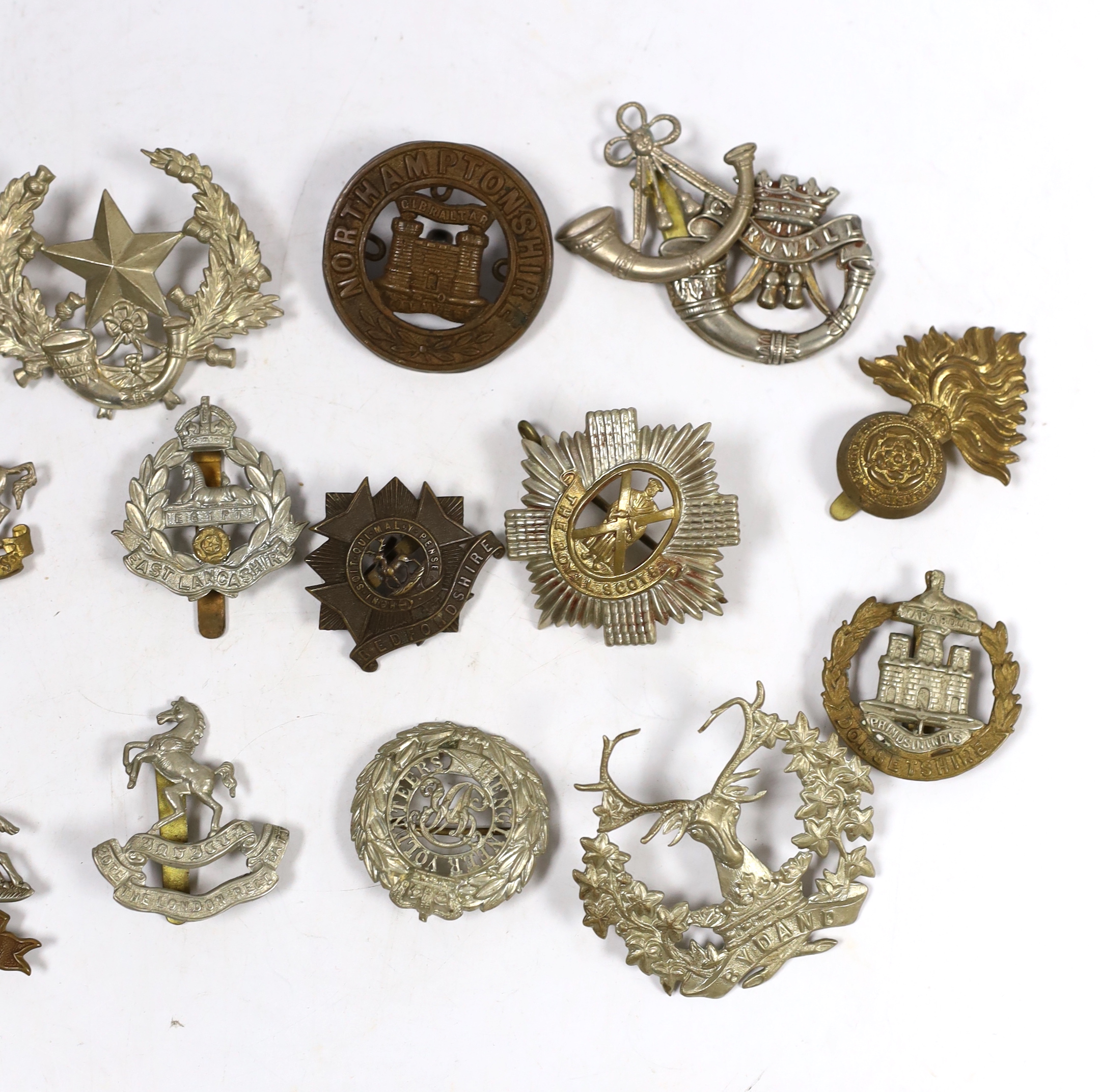 Twenty military cap badges including Bedfordshire, West Yorkshire, Cornwall, East Lancashire, the Royal Scots, the Royal Sussex, Northamptonshire, Lincolnshire, East Surrey, Dorsetshire, Engineer Volunteers, etc.